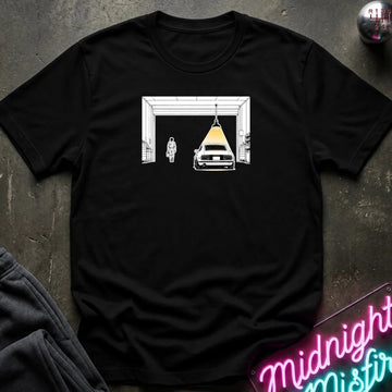 A black short-sleeve crewneck t-shirt laid flat on a dark textured surface. The shirt features a graphic print of a white classic car parked inside a white garage door frame, with a yellow spotlight shining down on it.