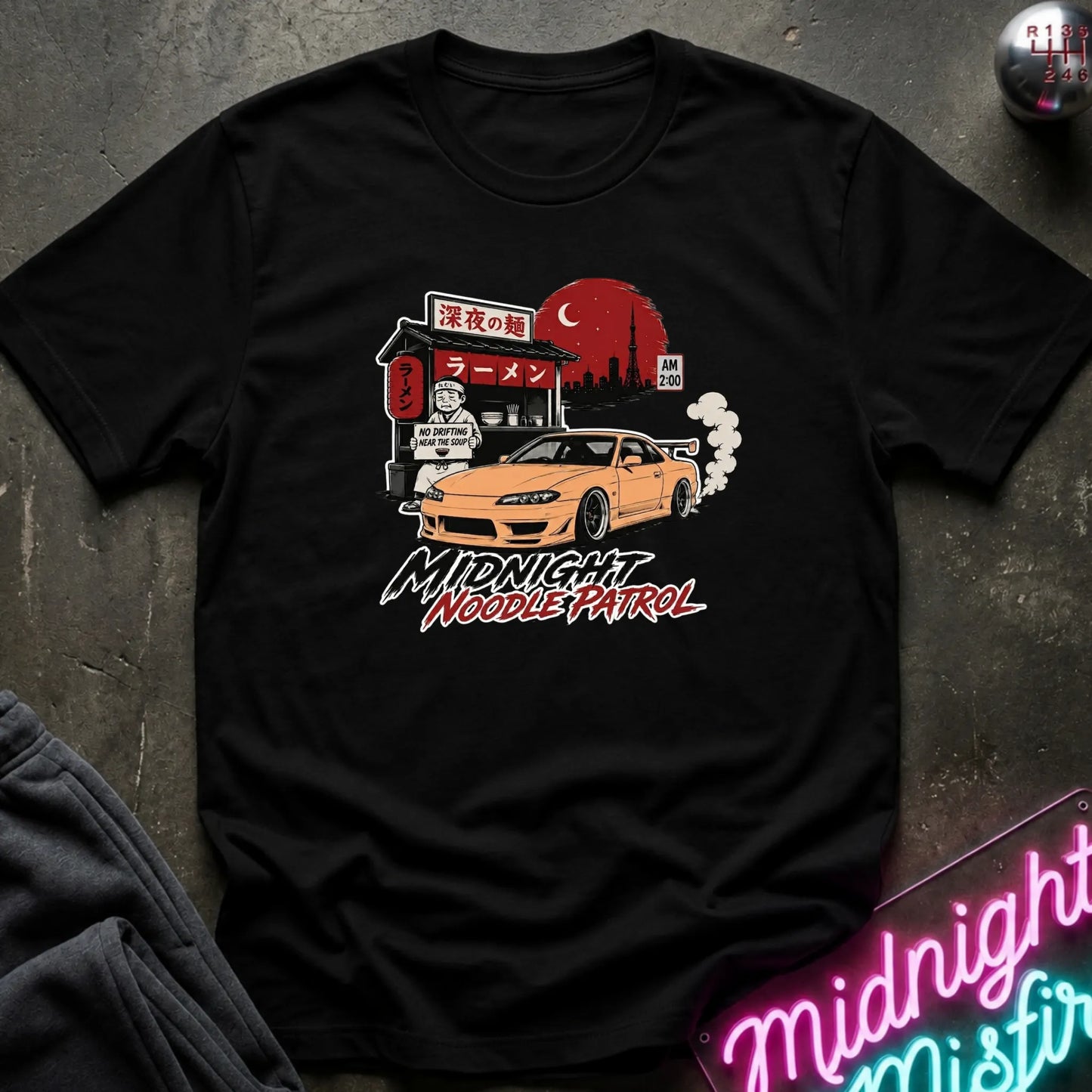 A black short-sleeve graphic t-shirt laid flat on a dark textured surface. The graphic features a yellow sports car in front of a ramen shop with a large red sun and the text 'Midnight Noodle Patrol'.