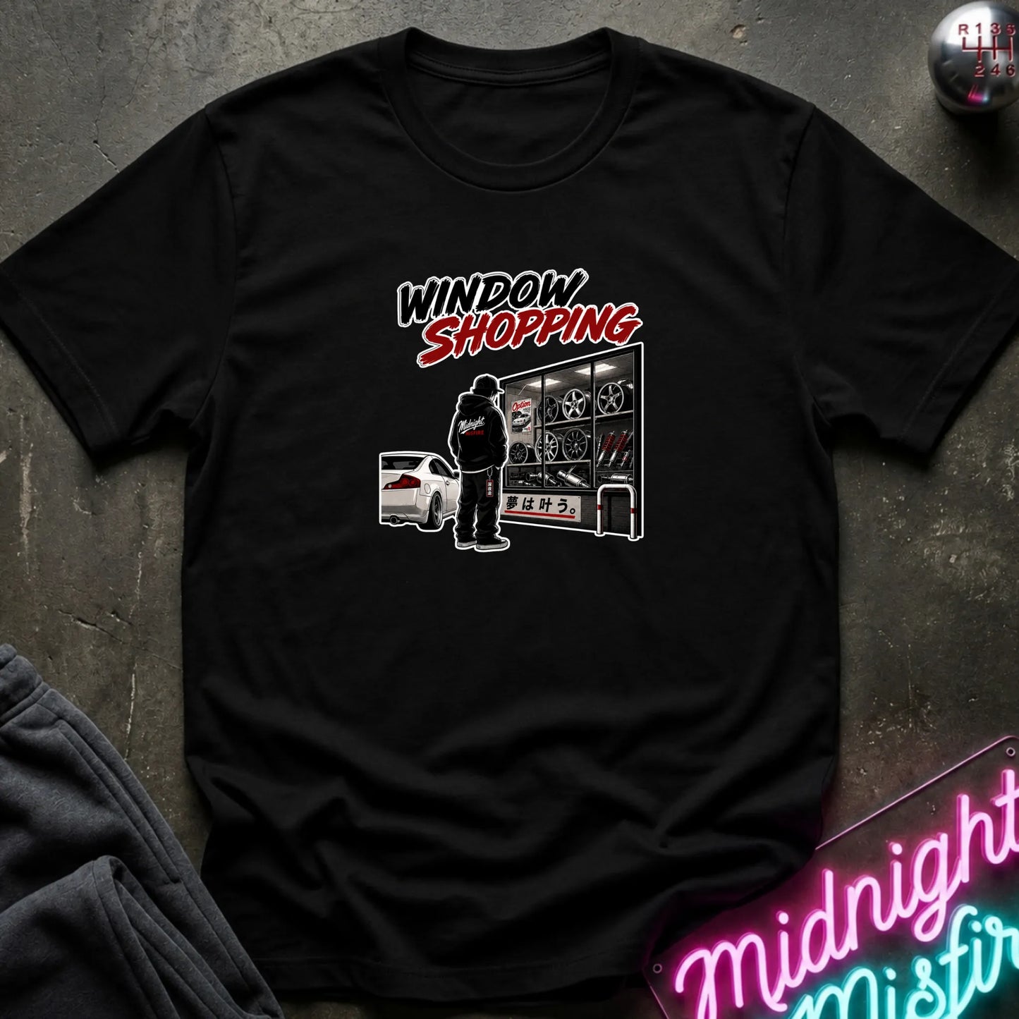 A black short-sleeved t-shirt  that has a cars parked and a person checking out windows of parts.