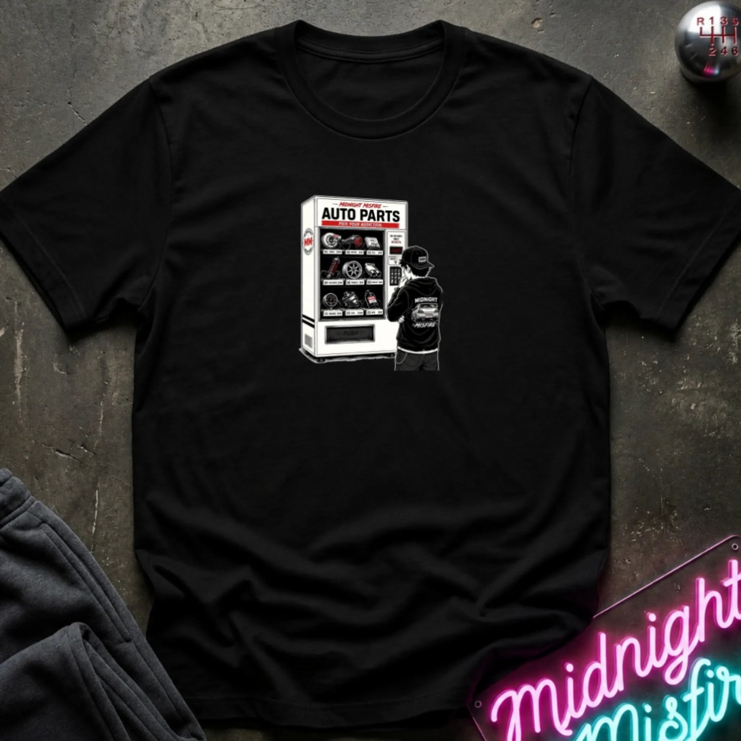 A black short-sleeve t-shirt laid flat on a dark textured surface, featuring a graphic print of a vending machine and owner deciding.
