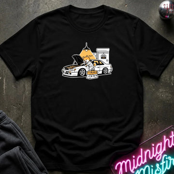 black graphic t-shirt laid flat on a dark textured surface. The graphic features a dog fixing a car.