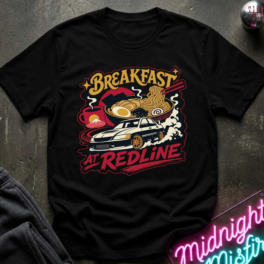 A black short-sleeve graphic T-shirt laid flat on a dark textured surface. The shirt features a colorful retro-style print with the words “Breakfast at Redline,” a white car, ramen noodles, an egg, and red abstract shapes.