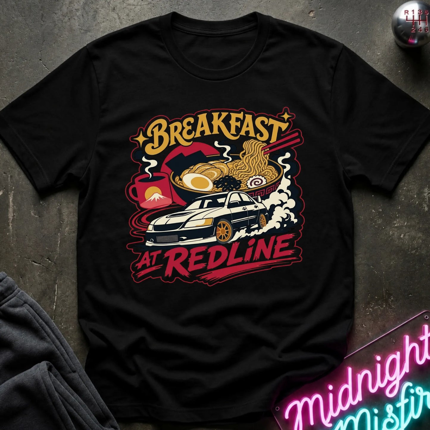 A black short-sleeve graphic T-shirt laid flat on a dark textured surface. The shirt features a colorful retro-style print with the words “Breakfast at Redline,” a white car, ramen noodles, an egg, and red abstract shapes.