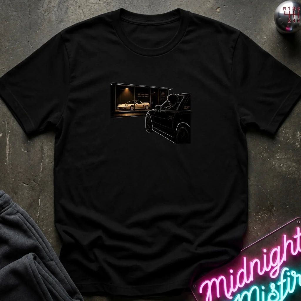A black short-sleeve crewneck t-shirt laid flat on a dark textured surface. The shirt features a graphic print on the chest showing a black car silhouette in the foreground and a white sports car inside a lit garage in the background.