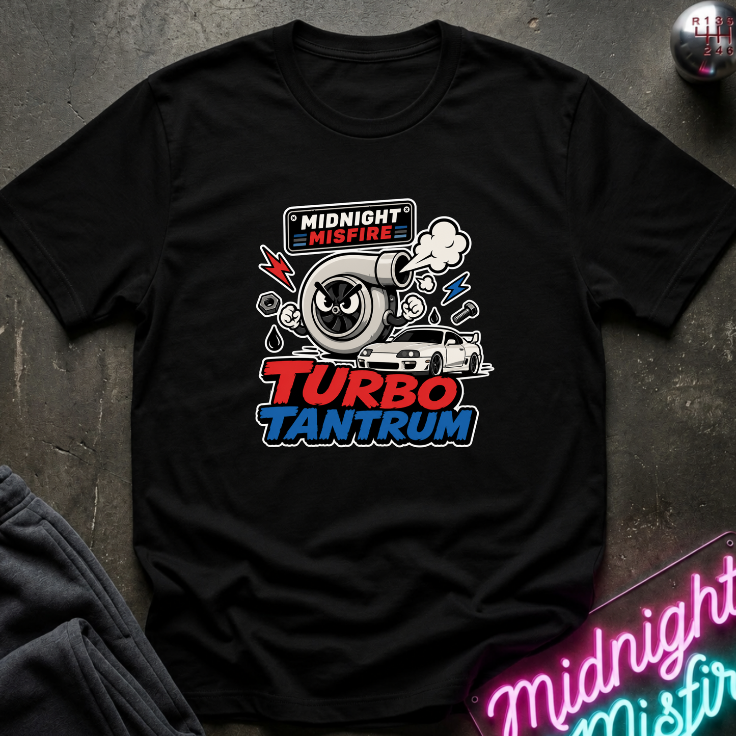 A black short-sleeve graphic t-shirt laid flat on a dark textured surface. The shirt features a colorful chest print with a cartoon turbocharger, a white car, and the text 'MIDNIGHT MISFIRE' and 'TURBO TANTRUM' in red, white, and blue.