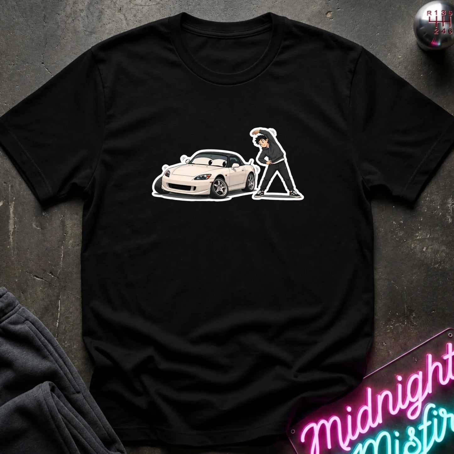 A black short-sleeve t-shirt laid flat on a dark textured surface, featuring a graphic print of a white sports car. A neon sign is partially visible in the bottom right corner.