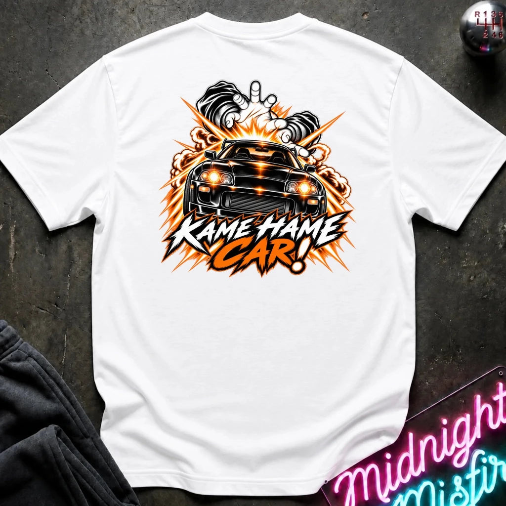 A white short-sleeve t-shirt laid flat on a dark textured surface, featuring a large graphic print of a black sports car with flames and the text 'KAME HAME CAR!' in orange and black. A small gear shift knob and neon sign are partially visible as props.