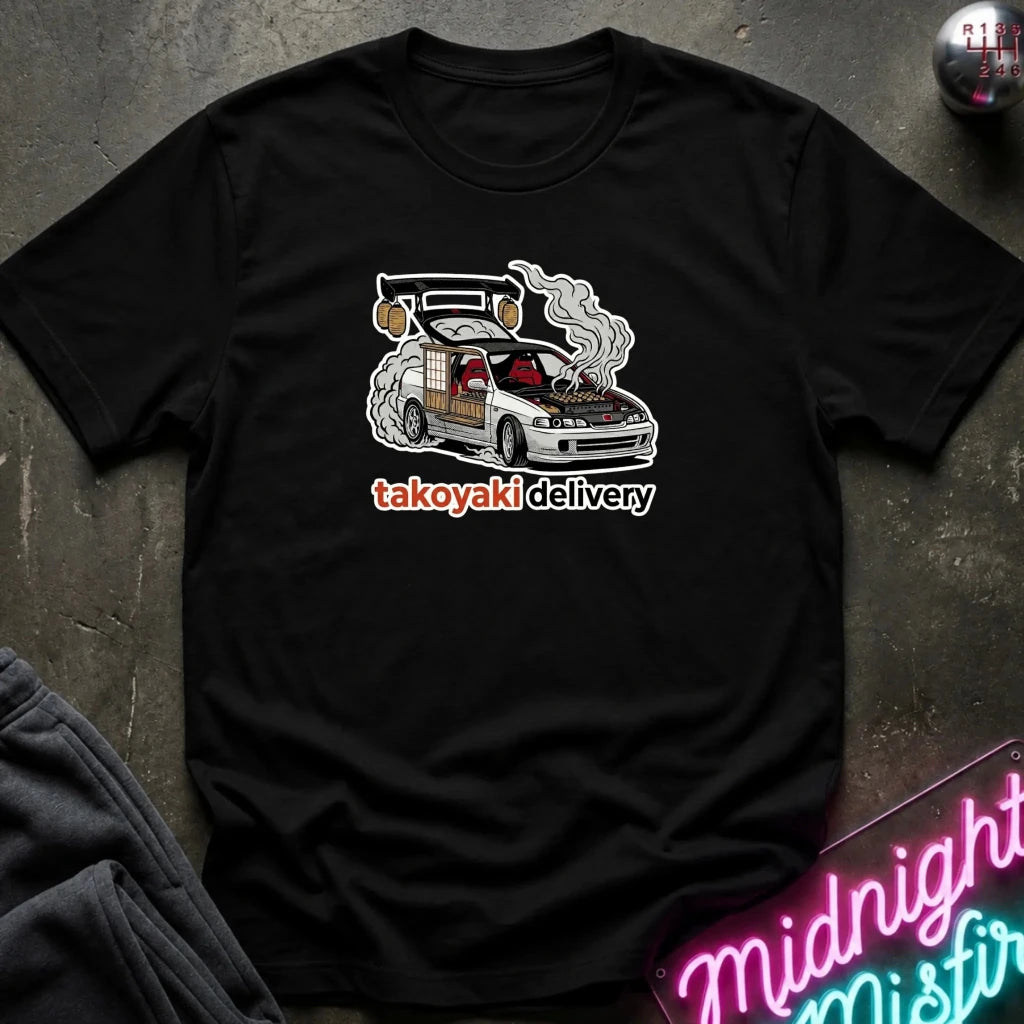 A black short-sleeve graphic t-shirt laid flat on a dark textured surface. The shirt features a graphic of a white car with a smoking engine and the text 'tskoyaki delivery' in white and orange.