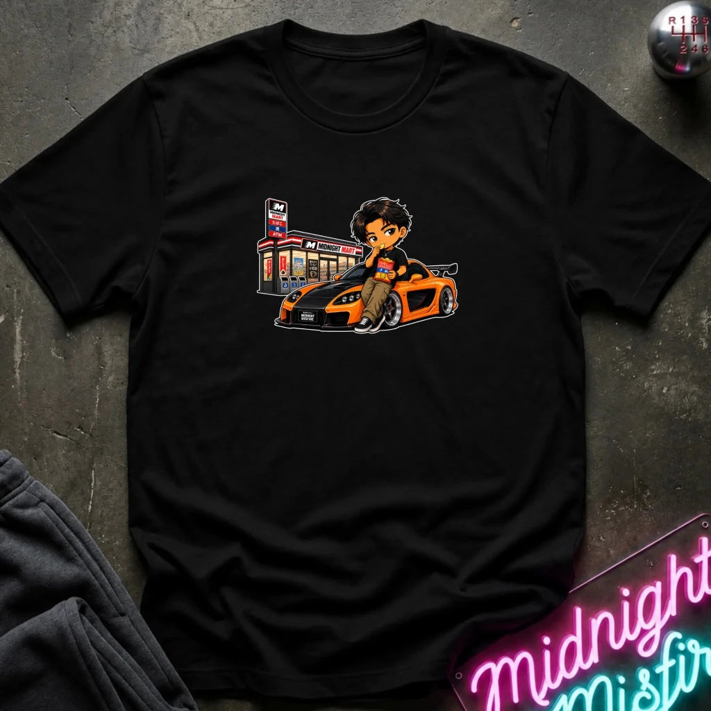 A black short-sleeve crewneck t-shirt laid flat on a dark textured surface, featuring a cartoon graphic of a man sitting on a orange and black sports car next to a store.