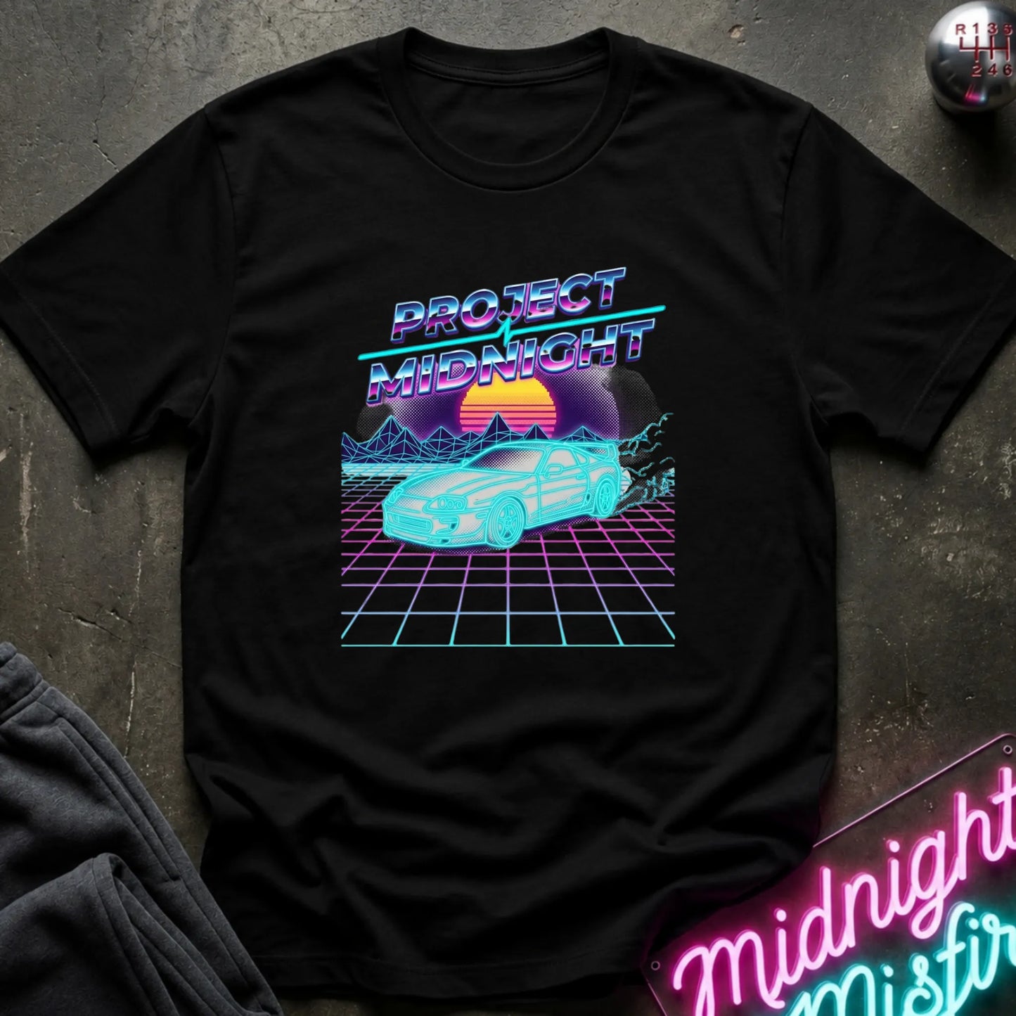 A black short-sleeve t-shirt with a retro 80s synthwave graphic reading 'PROJECT MIDNIGHT' above a glowing cyan car on a purple grid with a sunset, laid flat on a dark textured surface with a neon sign and folded gray fabric nearby.