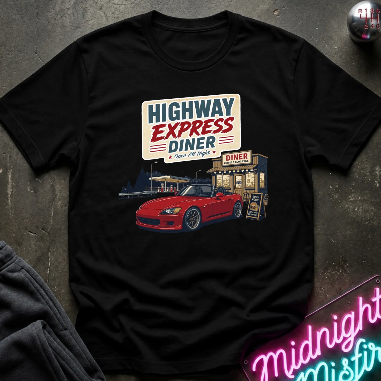 A black short-sleeve graphic t-shirt laid flat on a dark textured surface. The shirt features a retro-style print of a red sports car parked outside a diner with a sign reading 'HIGHWAY EXPRESS DINER'.