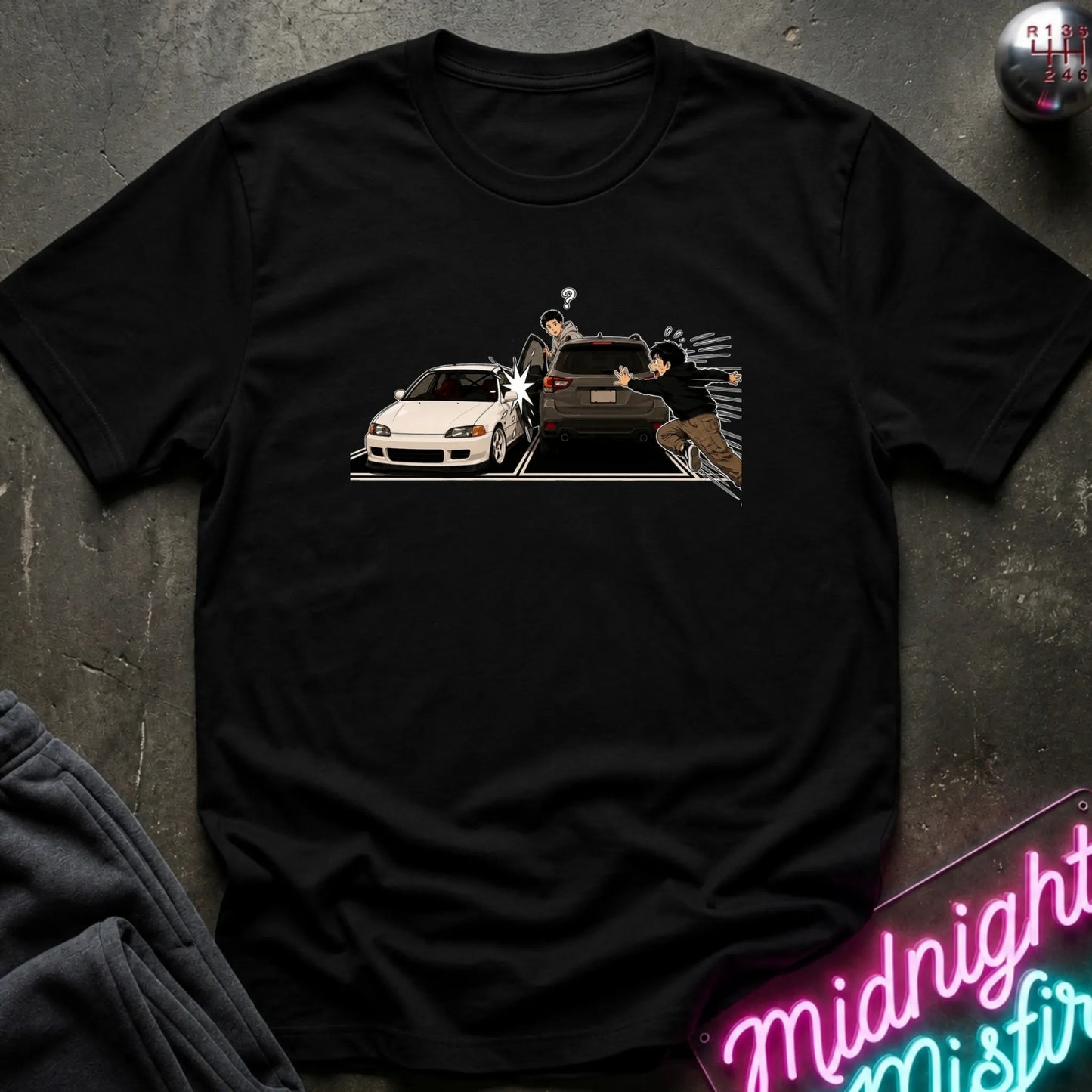 A black short-sleeve crewneck t-shirt laid flat on a dark textured surface. The center of the shirt features a graphic print depicting a white sports car and a dark SUV with two characters in a street-art style.