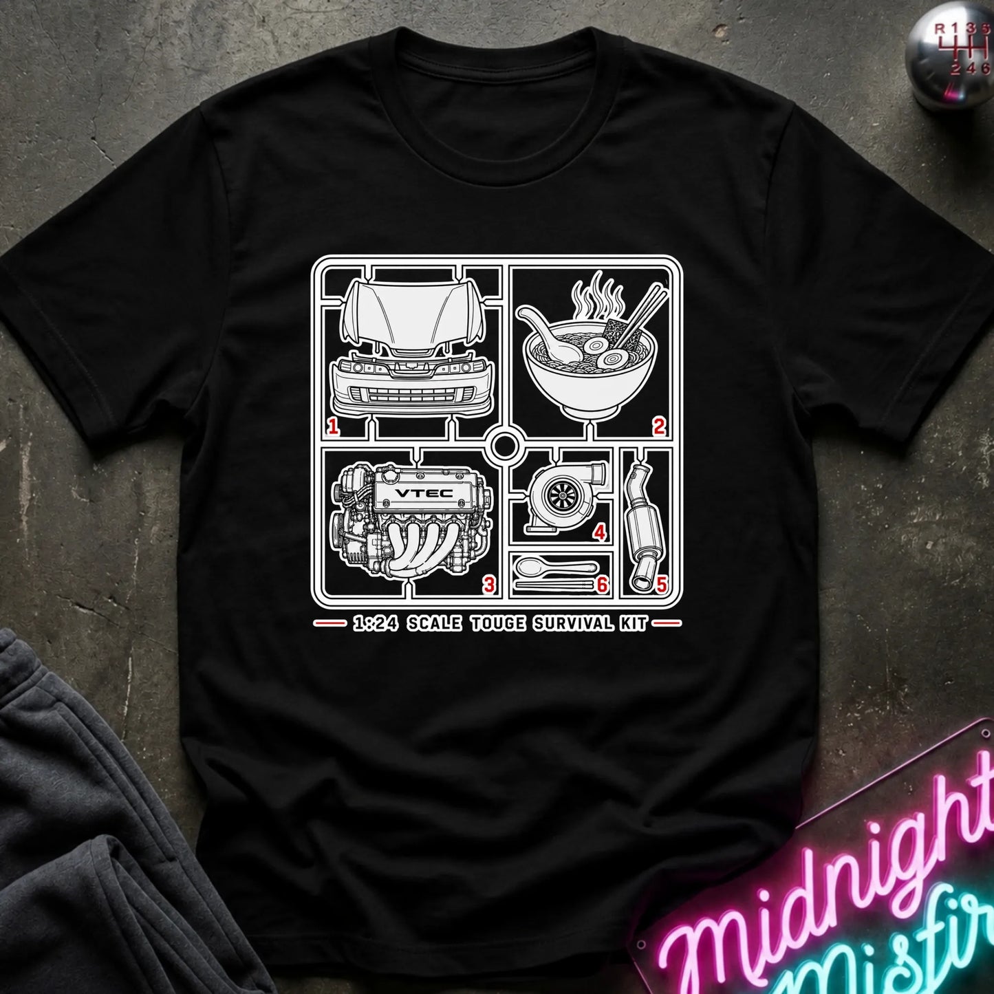A black short-sleeve graphic T-shirt laid flat on a dark textured surface. The shirt features a black line-art schematic of a survival kit with numbered illustrations and the text '1:24 SCALE TOUGHE SURVIVAL KIT' printed on the chest.