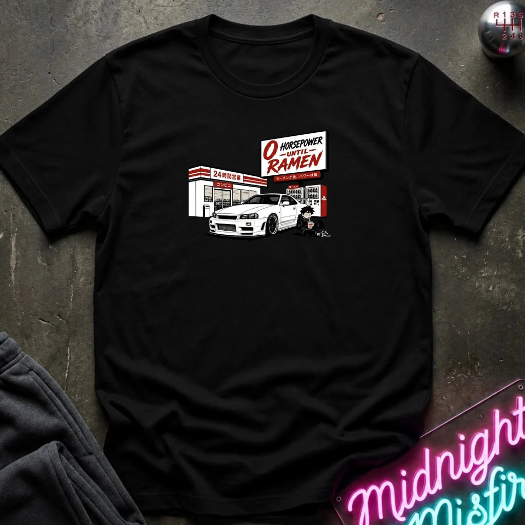 A black crewneck t-shirt laid flat on a dark textured surface, featuring a graphic print of a white sports car parked in front of a konbini.