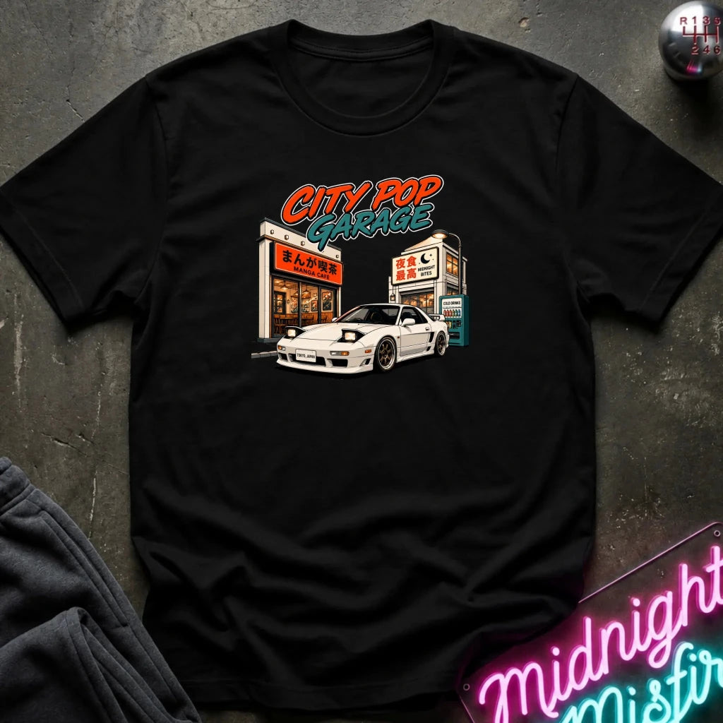 A black short-sleeve graphic t-shirt laid flat on a dark textured surface. The graphic features a white sports car parked in front of a garage with the text 'CITY POP GARAGE' in orange and teal.