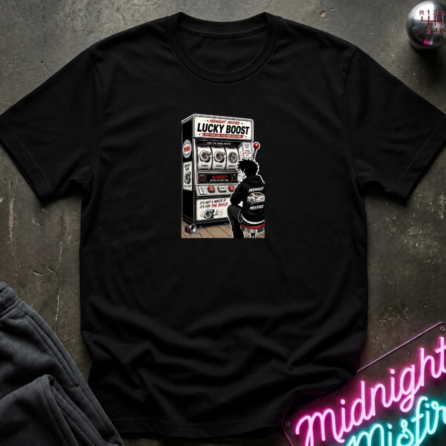 A black  short-sleeve t-shirt with a retro-style graphic of a slot machine, displayed flat on a dark textured surface with a neon sign and sweatpants partially visible.
