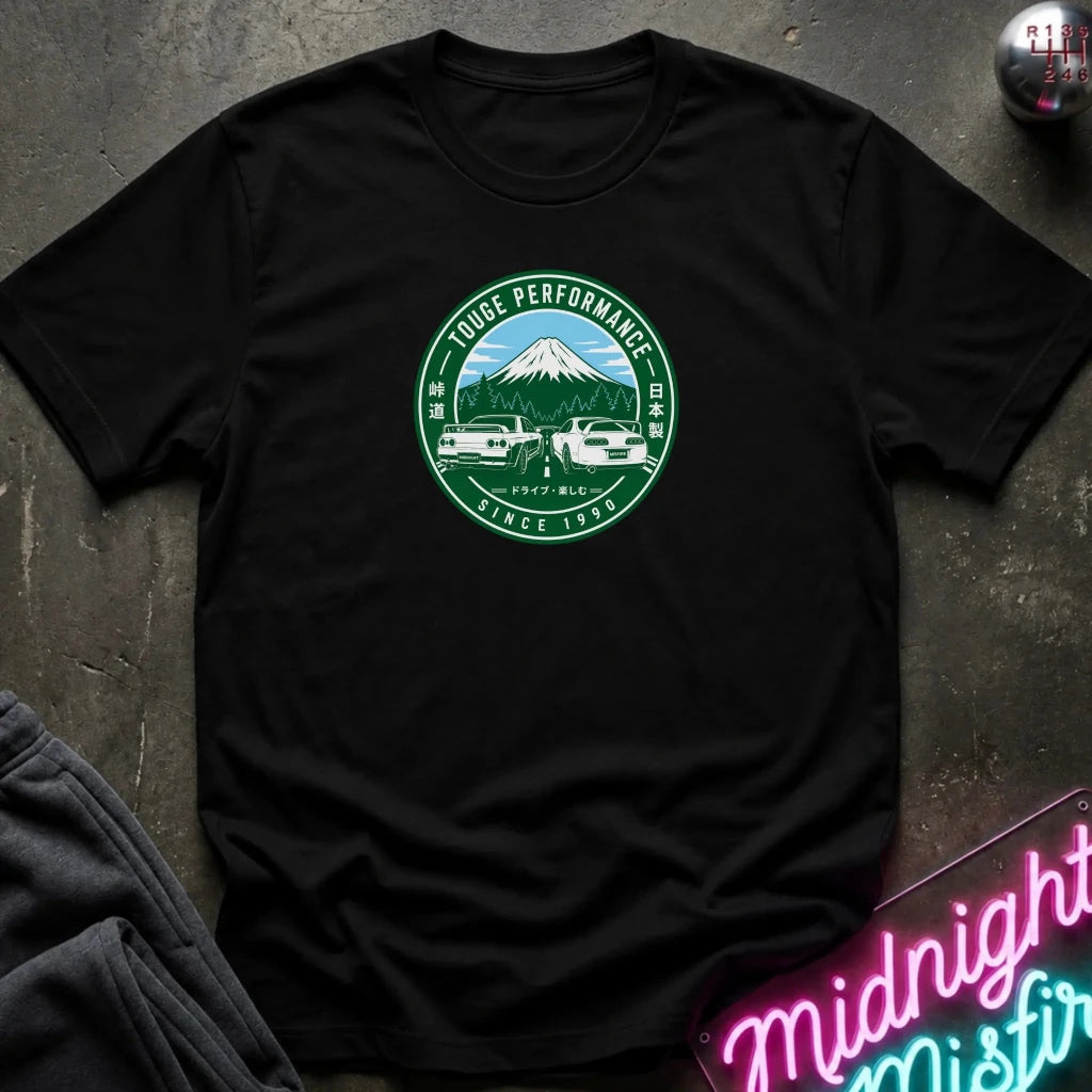 A black short-sleeve t-shirt laid flat on a dark textured surface, featuring a circular green and white graphic on the chest with a mountain, two cars, and the text 