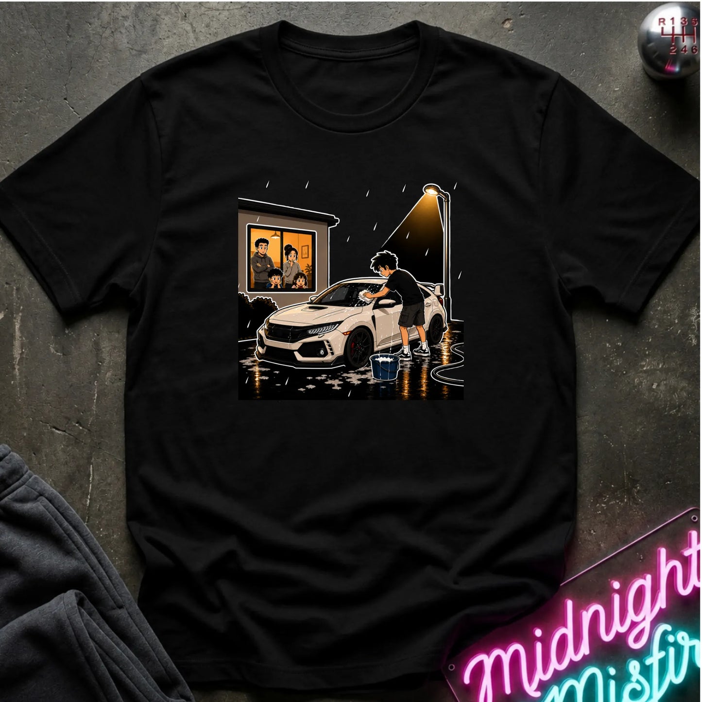 A black short-sleeve graphic t-shirt laid flat on a dark textured surface. The graphic shows a person washing a white sports car in the rain, with a window showing  people inside and a glowing streetlight.