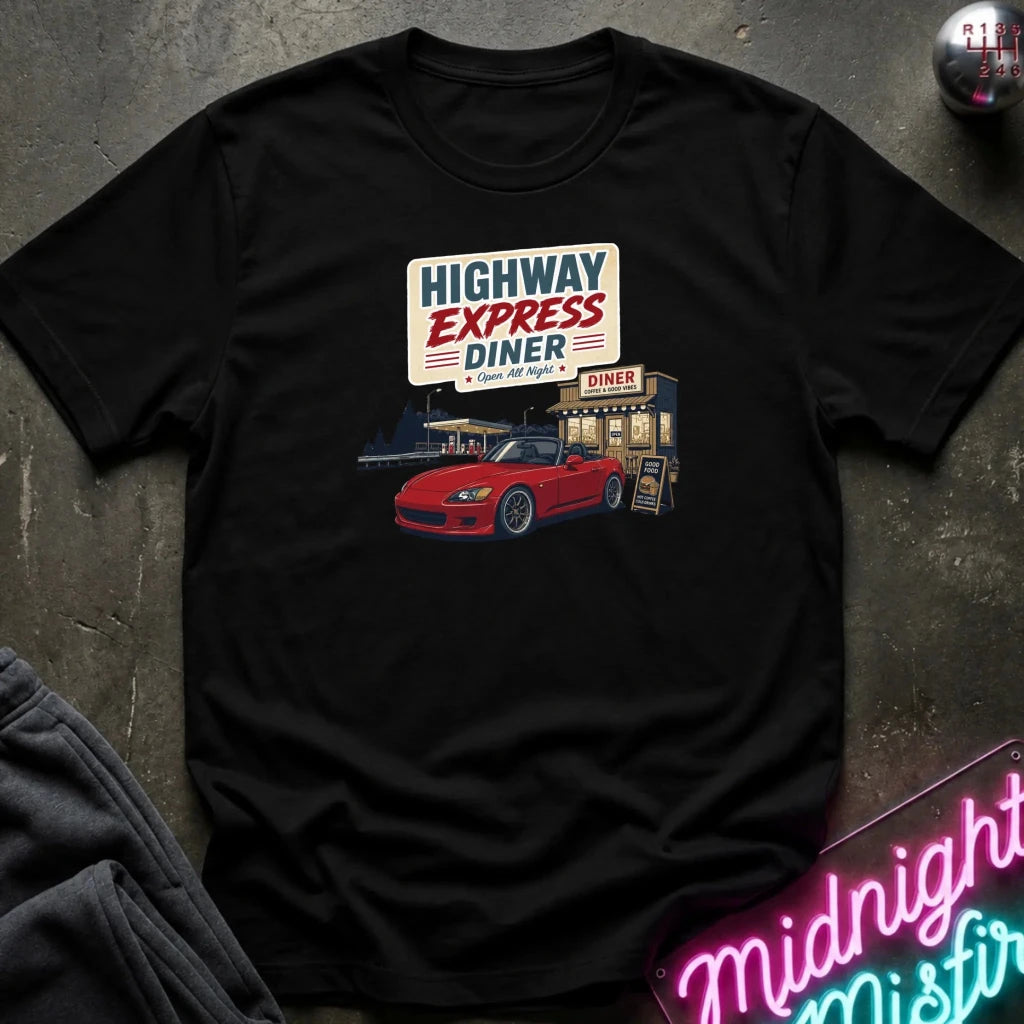 A black short-sleeve graphic t-shirt laid flat on a dark textured surface. The shirt features a retro-style print of a 'Highway Express Diner' with a red sports car and a neon sign in the foreground.