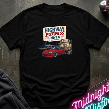 A black short-sleeve graphic t-shirt laid flat on a dark textured surface. The shirt features a retro-style print of a 'Highway Express Diner' with a red sports car and a neon sign in the foreground.
