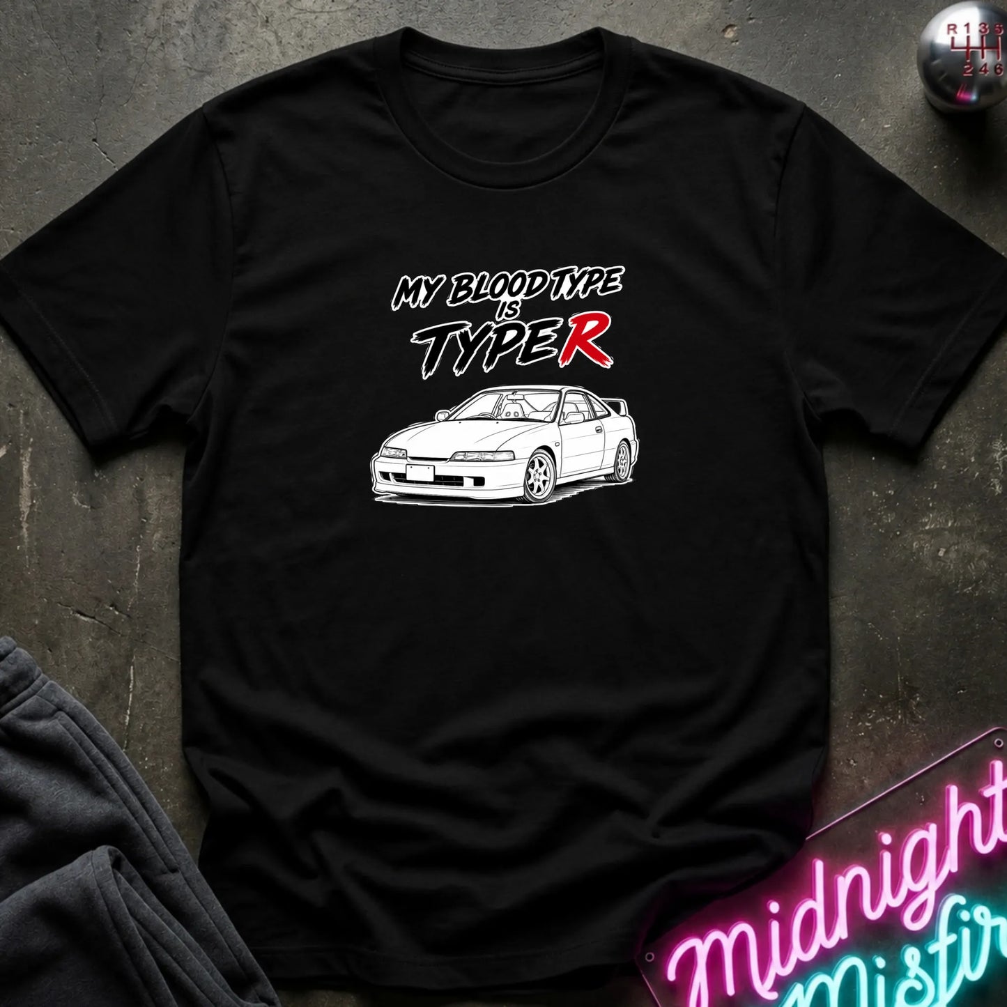 A black short-sleeve graphic T-shirt laid flat on a dark textured surface. The shirt features a white line drawing of a sports car with the text 'MY BLOODTYPE IS TYPER' in white and red lettering.