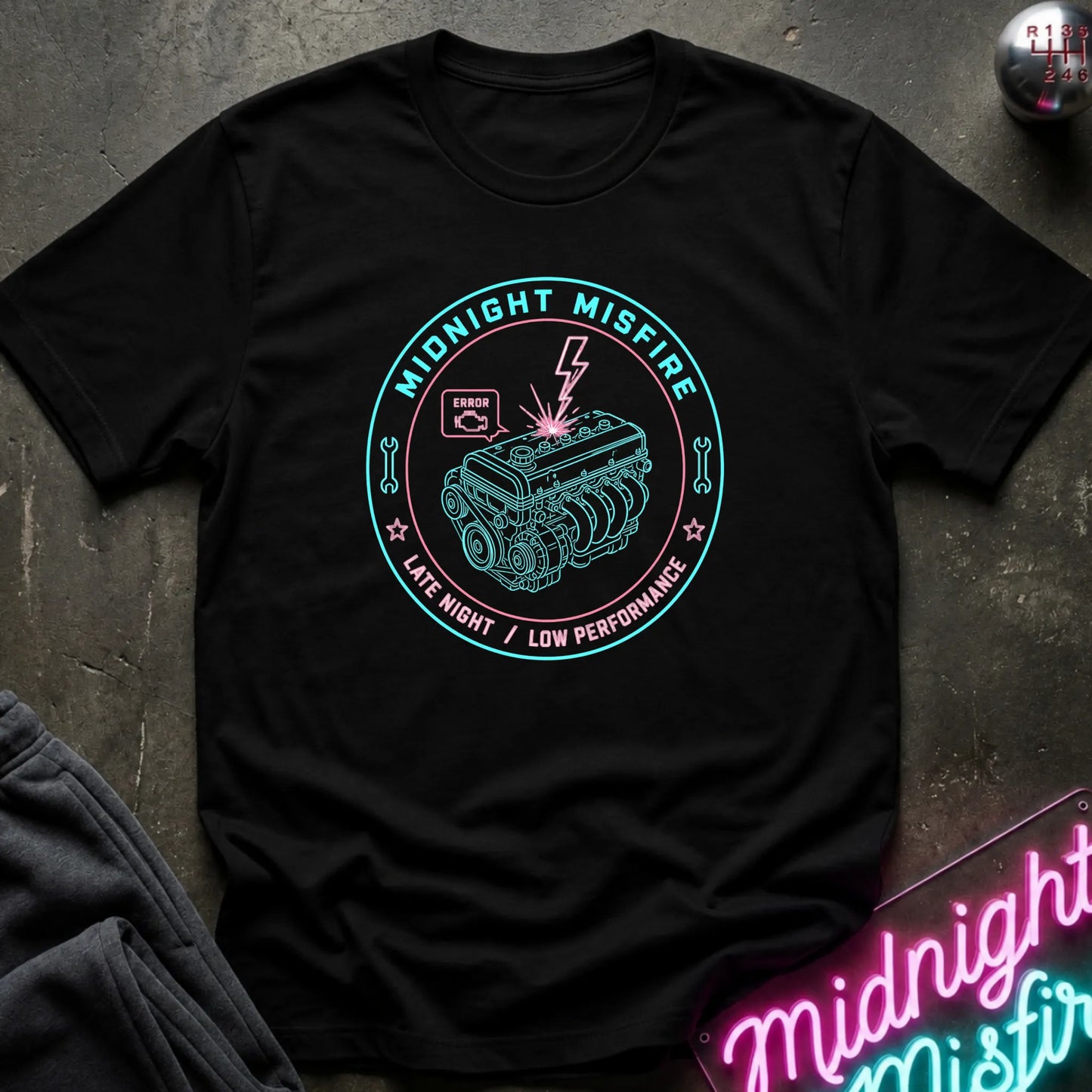 A black short-sleeve graphic T-shirt laid flat on a dark textured surface. The shirt features a circular teal and pink graphic of a car engine with the words 'MIDNIGHT MISFIRE' and 'LATE NIGHT / LOW PERFORMANCE.