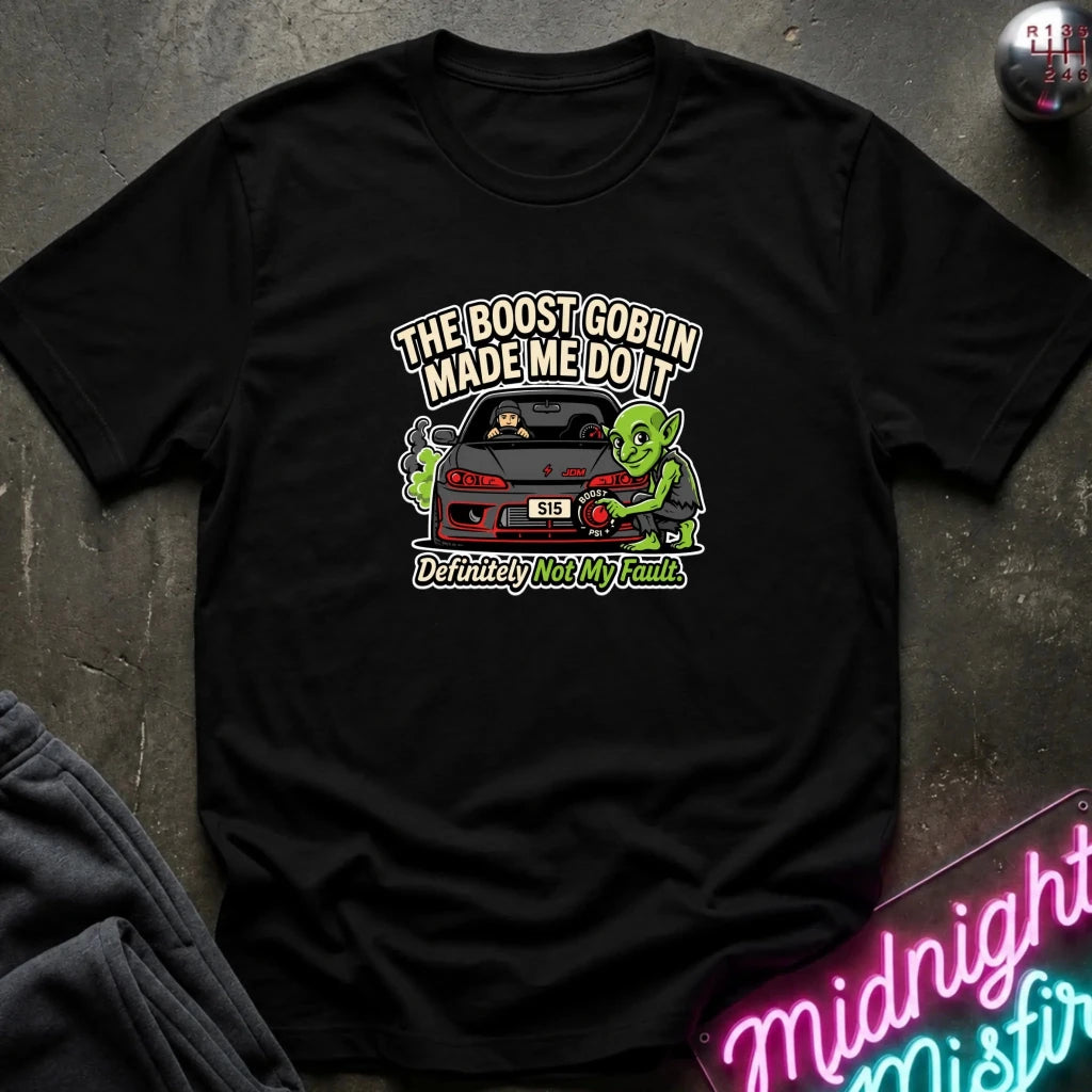 A black short-sleeve graphic t-shirt laid flat on a dark textured surface. The graphic features a green cartoon character next to a red sports car with the text 'THE BOOST GOBLIN MADE ME DO IT' above and 'Definitely Not My Fault.