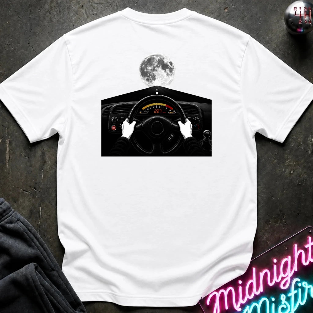 A white short-sleeve t-shirt laid flat on a dark textured surface, featuring a graphic print of a driver's hands on a steering wheel with a full moon above. A neon sign and dark pants are partially visible as props.