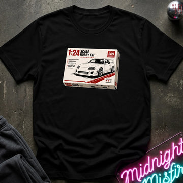 A black short-sleeve t-shirt laid flat on a dark textured surface, featuring a printed graphic of a 1:24 scale model hobby kit box for a white sports car. The shirt is styled with a gray sweatshirt corner and a pink neon sign in the background.