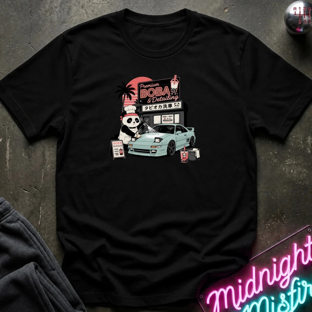 A black short-sleeve graphic t-shirt laid flat on a dark textured surface. The graphic features a cartoon panda washinga light blue car