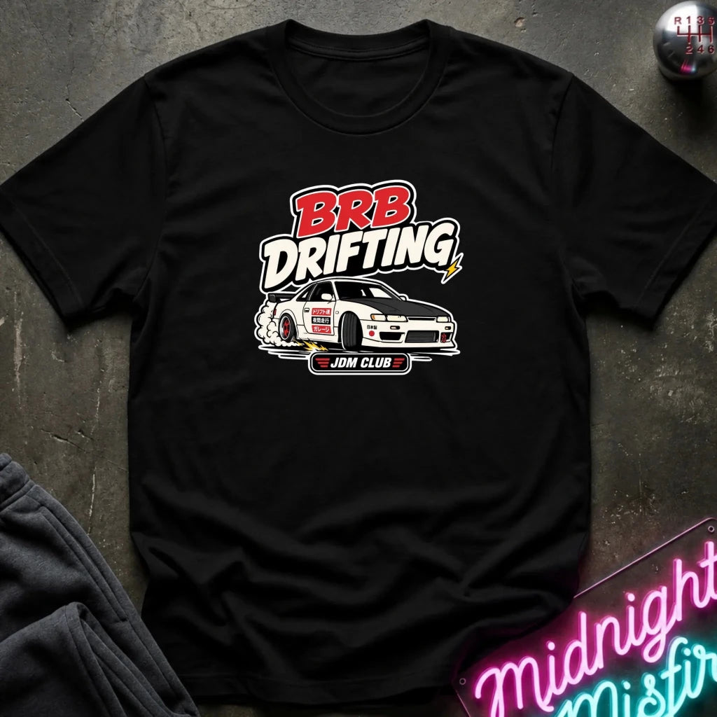 A black short-sleeve graphic T-shirt laid flat on a dark textured surface. The shirt features a large front print with the words “BRB Drifting” in red and white