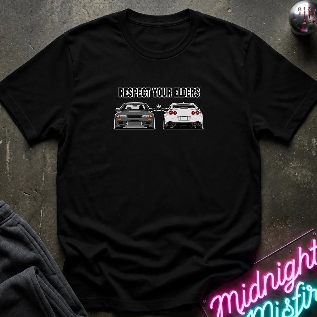 A black short-sleeve graphic t-shirt laid flat on a dark textured surface. The shirt features a white graphic print with the text 'RESPECT YOUR ELDERS' above two sports cars, one gray and one white.