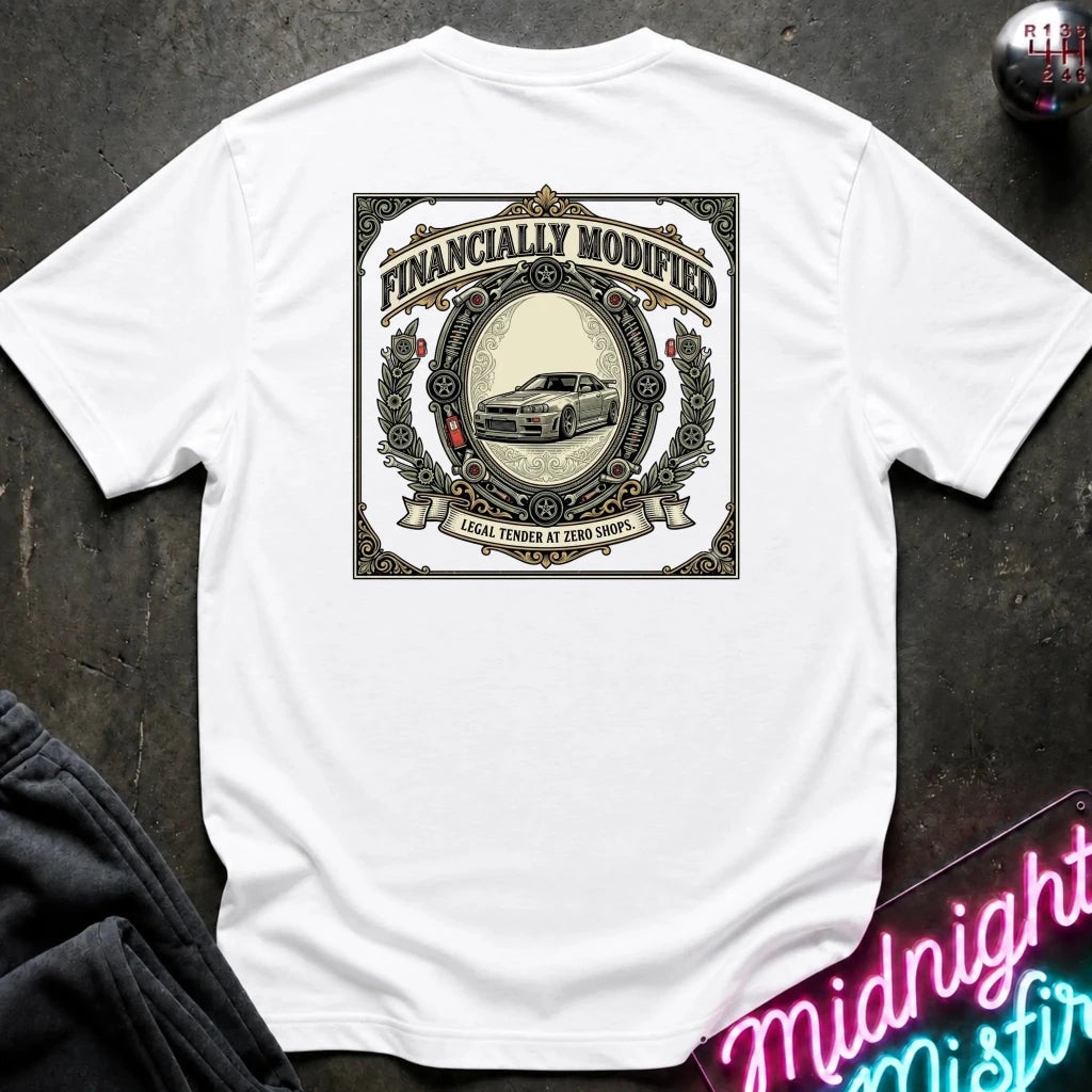 A white short-sleeve t-shirt laid flat on a dark textured surface, featuring a large vintage-style graphic on the back with the words 'FINANCALLY MODIFIED' and an illustration of a car. A neon sign and dark clothing are partially visible around the edges as styling props.