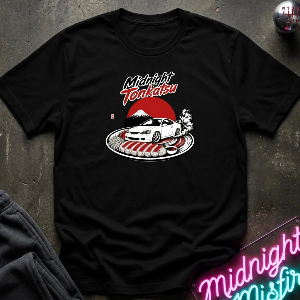 A black short-sleeve graphic t-shirt laid flat on a dark textured surface. The graphic features the words 'Midnight Tenkasu' in stylized white and red lettering above a white sports car, a red sun, and a Japanese mountain scene.