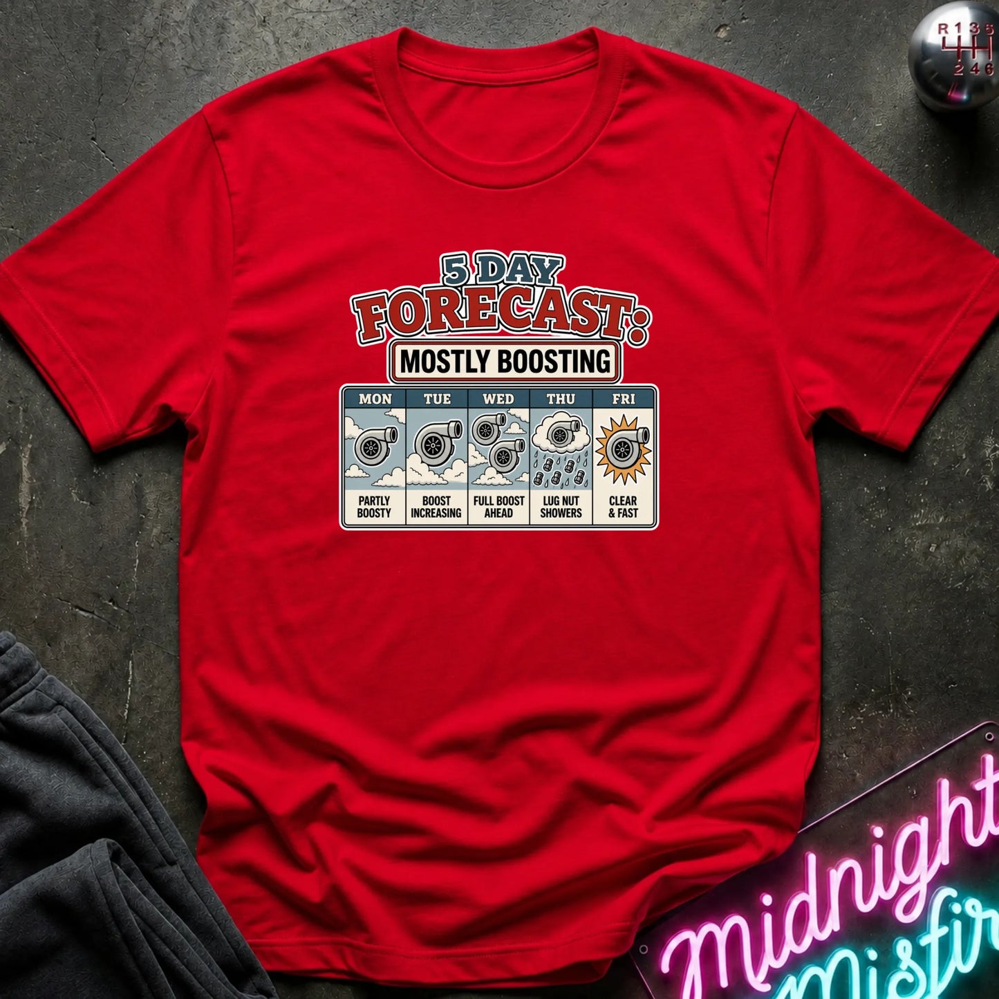a red short-sleeve graphic T-shirt laid flat on a dark textured surface. The shirt features a colorful '3 Day Forecast: Mostly Boosting' print with weather icons and text for Monday through Friday.