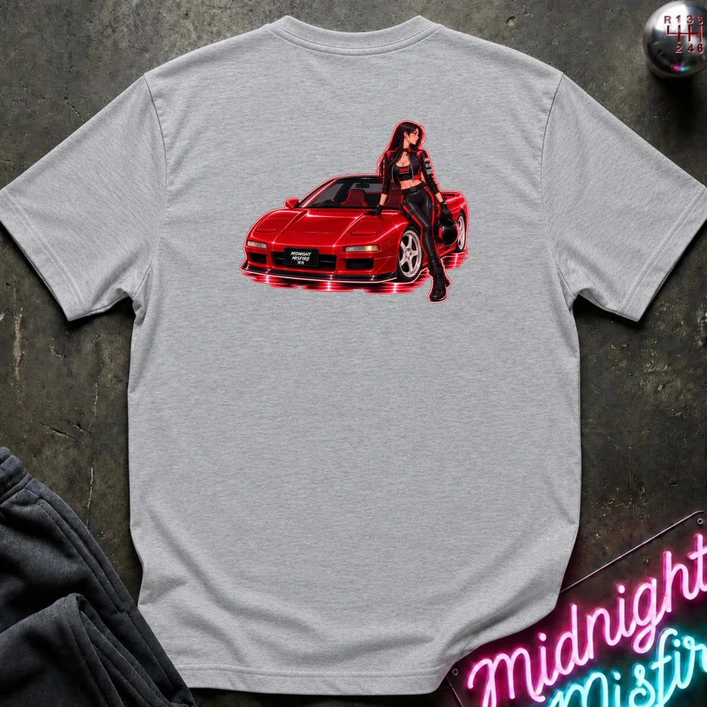 A light gray crewneck t-shirt laid flat on a dark textured surface, featuring a graphic print of a red sports car with a woman in black posing on top. A neon sign and dark sweatpants are partially visible in the corners.