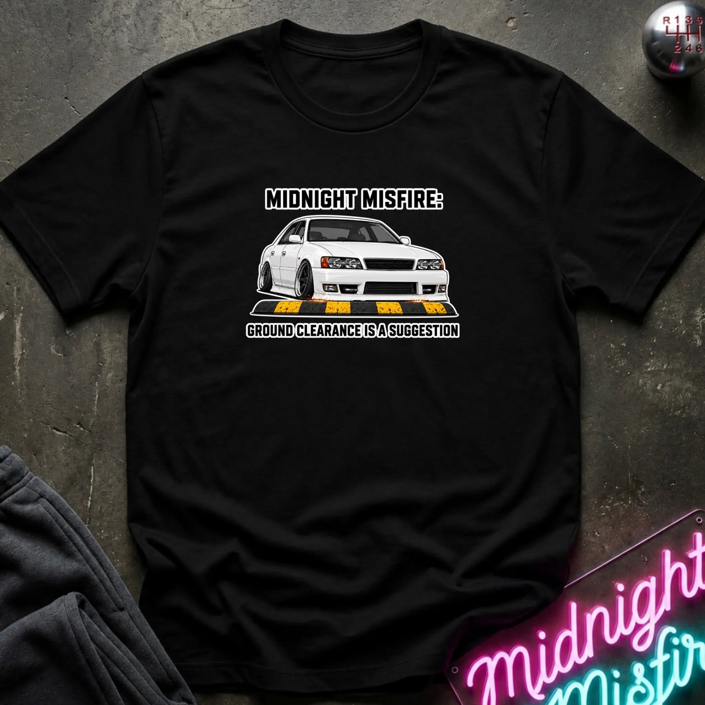 A black short-sleeve graphic t-shirt laid flat on a dark textured surface. The shirt features a white car graphic with yellow road markings and the text 'MIDNIGHT MISFARE' and 'GROUND CLEARANCE IS A SUGGESTION'.