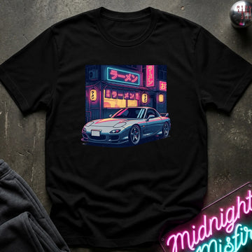 A black short-sleeve t-shirt laid flat on a dark textured surface, featuring a colorful graphic print of a blue sports car parked in front of a ramen shop with neon signs. A gray sweatshirt corner and a neon sign are partially visible as styled props.
