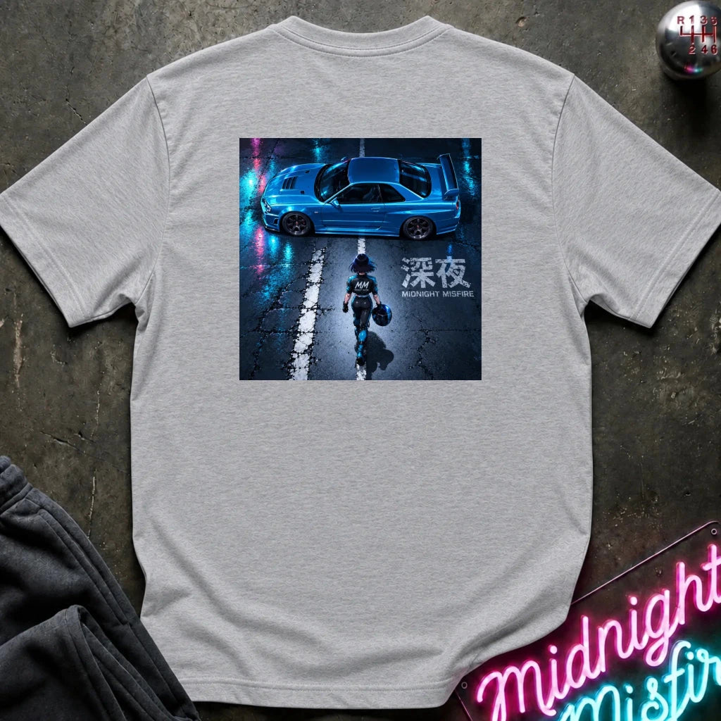 A light gray crewneck t-shirt laid flat on a dark textured surface, featuring a large square graphic print on the back with a blue car and a figure in a neon-lit scene. The shirt is styled with a dark sweatshirt in the corner and a neon sign in the foreground.