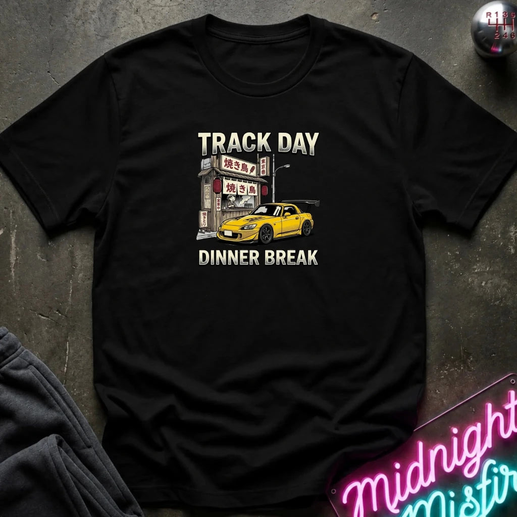 A black short-sleeve graphic T-shirt laid flat on a dark textured surface. The shirt features a printed design with the words 'TRACK DAY DINNER BREAK' and an illustration of a yellow sports car parked in front of a Japanese-style restaurant.