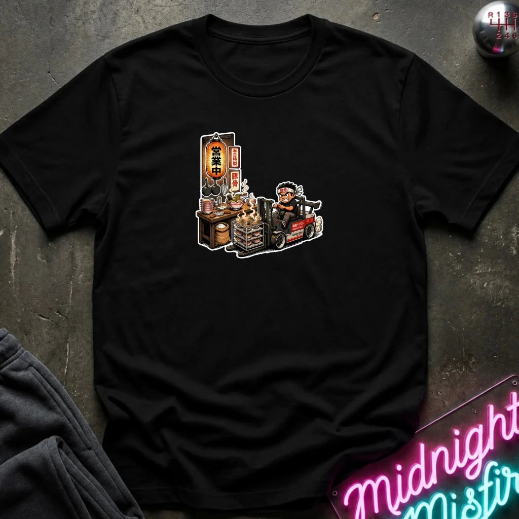 A  black short-sleeve t-shirt with a cartoon graphic of a street food vendor on a forklift, laid flat on a dark textured surface with a neon sign and folded sweatpants nearby.