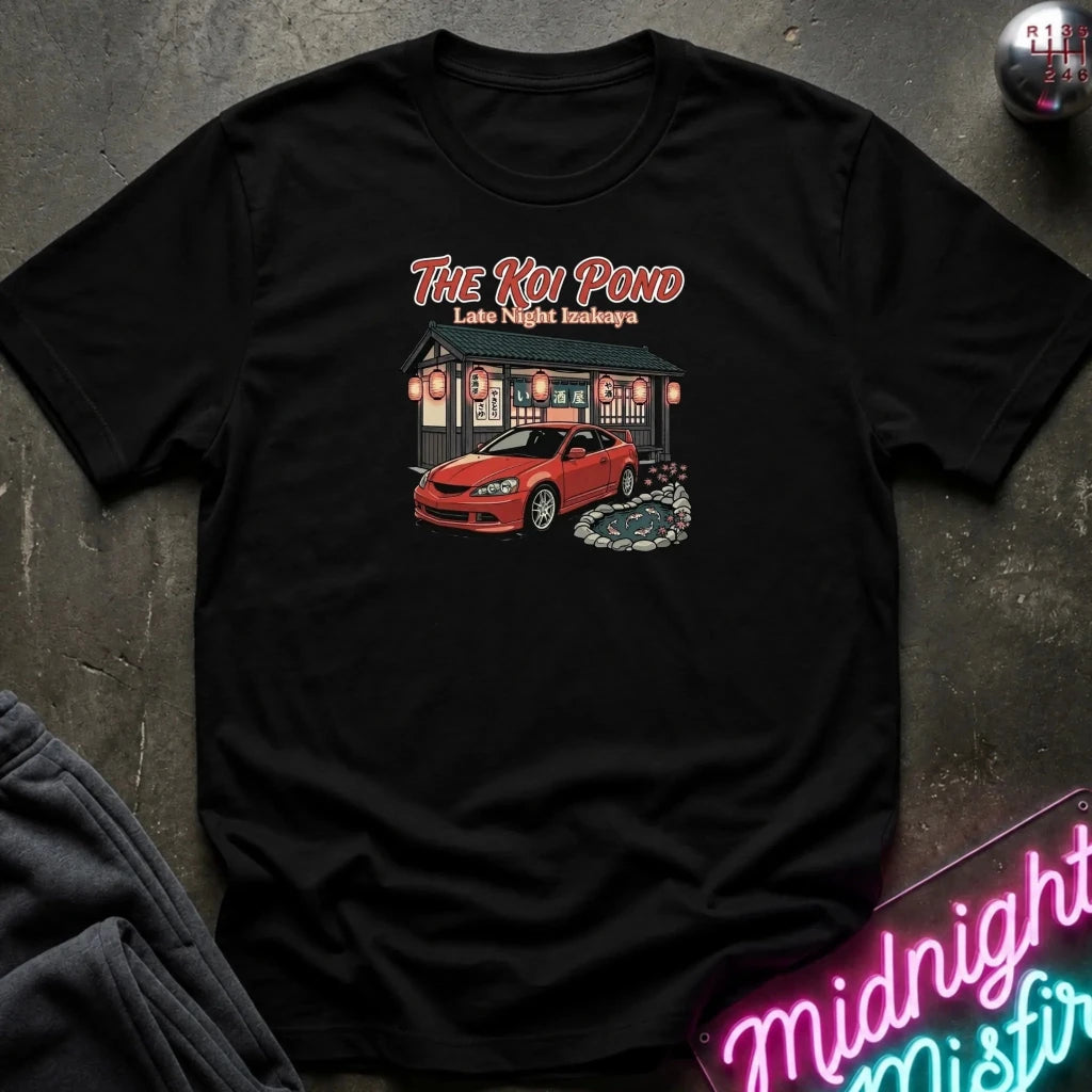 A black short-sleeve graphic t-shirt laid flat on a dark textured surface. The graphic features the text 'THE KOI POND Late Night Hamburger' above an illustration of a red sports car parked in front of a restaurant with red lanterns and a koi pond.