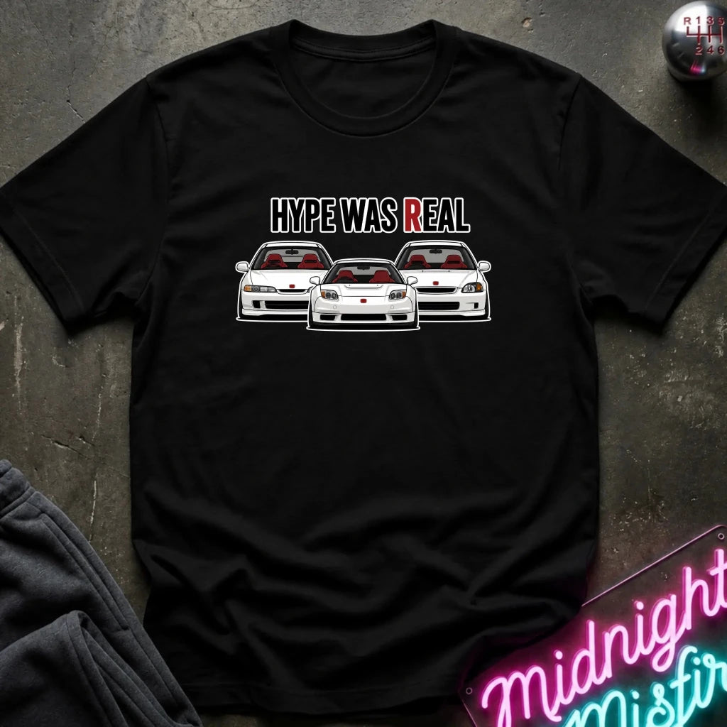A black short-sleeve graphic t-shirt laid flat on a dark textured surface. The shirt features a graphic of three white sports cars with red interiors and the text 'HYPE WAS REAL' in white and red.