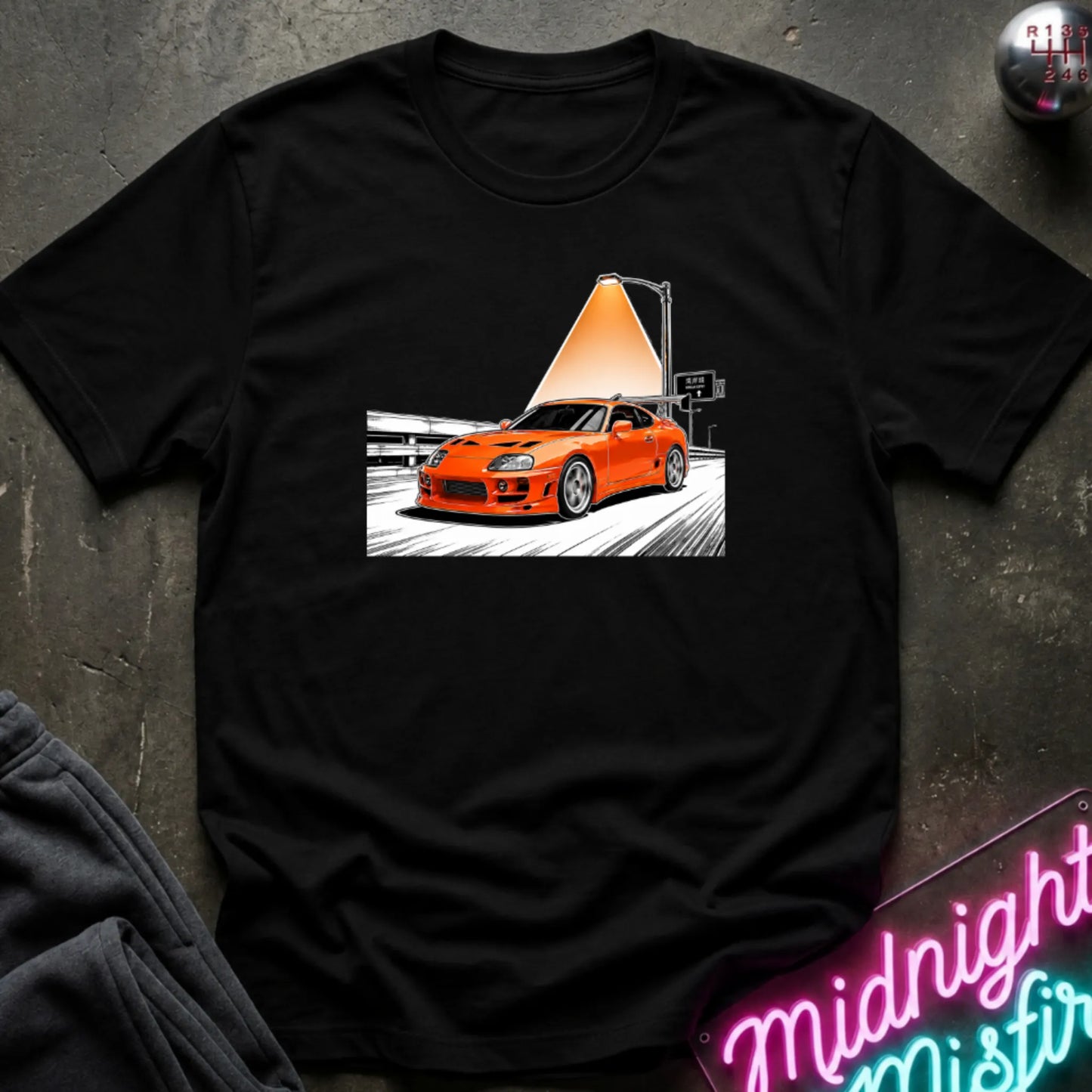 A black short-sleeve t-shirt laid flat on a dark textured surface, featuring a graphic print of an orange sports car speeding on highway.