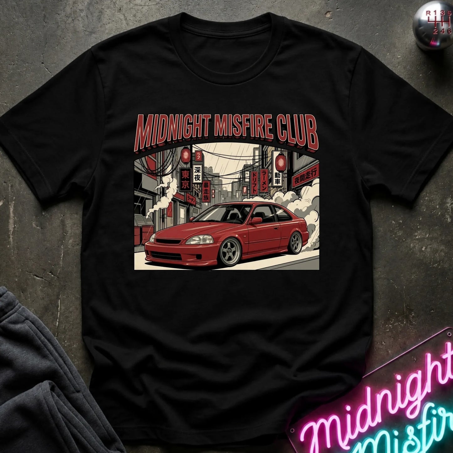 A black short-sleeve graphic t-shirt laid flat on a dark textured surface. The shirt features a central graphic of a red coupe car in a stylized city street with the text 'MIDNIGHT MISFIRE CLUB' in red above it.