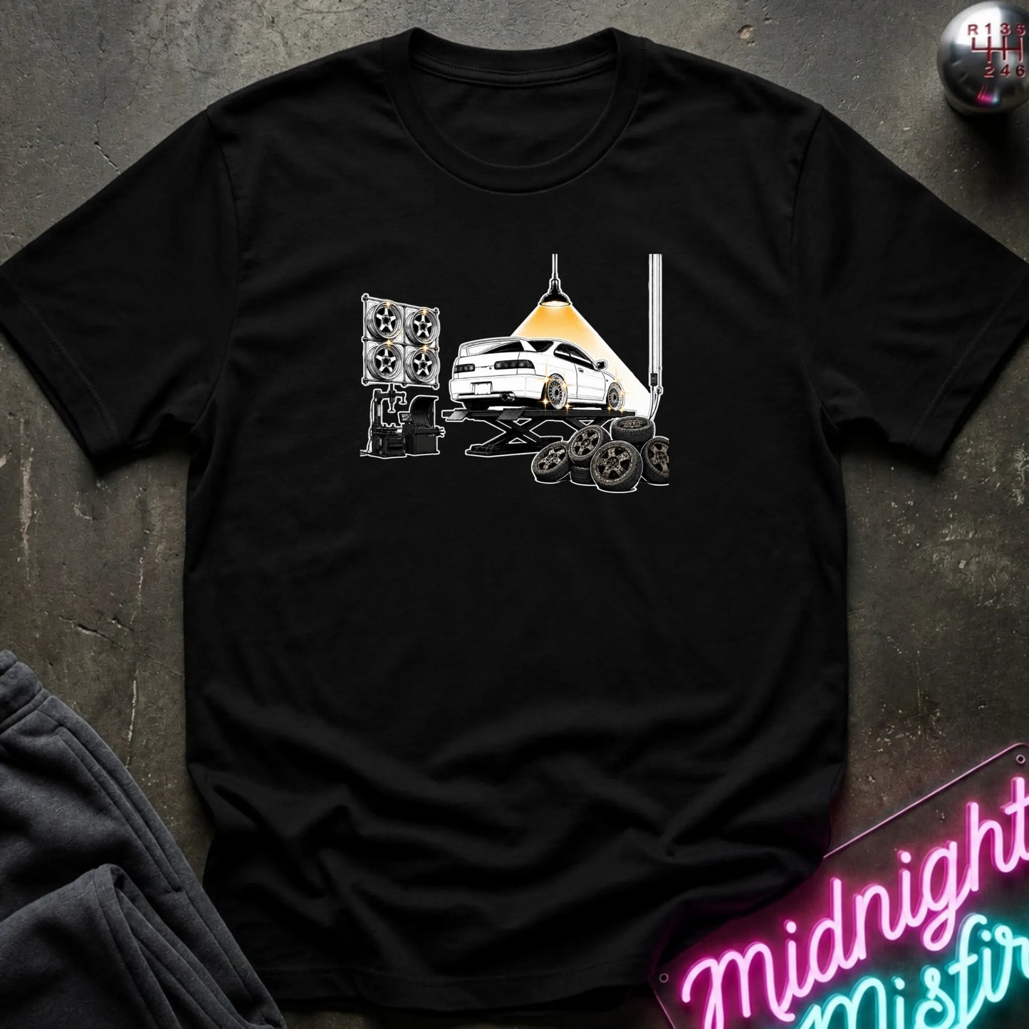 A black short-sleeve crewneck t-shirt laid flat on a dark textured surface. The shirt features a graphic print of a white car on a hydraulic lift with new wheels.
