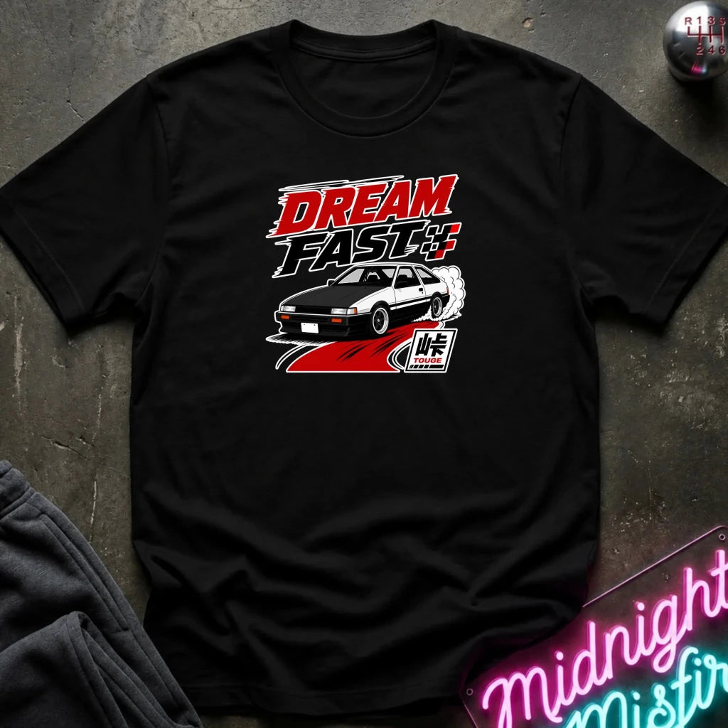 A black short-sleeve graphic T-shirt laid flat on a dark textured surface. The shirt features a red, black, and white 'DREAM FAST' graphic with a car illustration.
