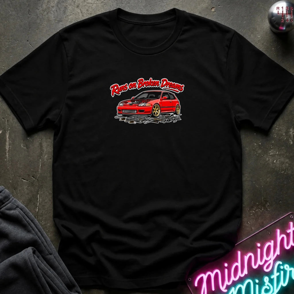 A black short-sleeve graphic t-shirt laid flat on a dark textured surface. The shirt features a red car illustration with the text 'Rust on Broken Dreams' in red and white.