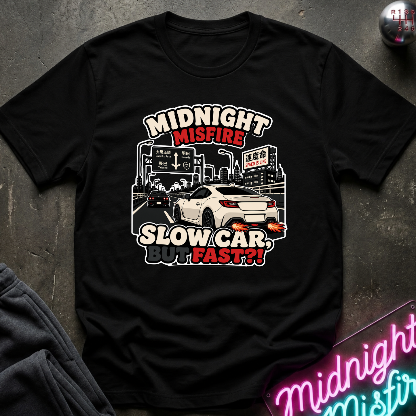 A black short-sleeve graphic T-shirt laid flat on a dark textured surface. The shirt features a white and red graphic print with the text 'MIDNIGHT MISFIRE SLOW CAR, FAST??' and an illustration of a white sports car in a city scene.