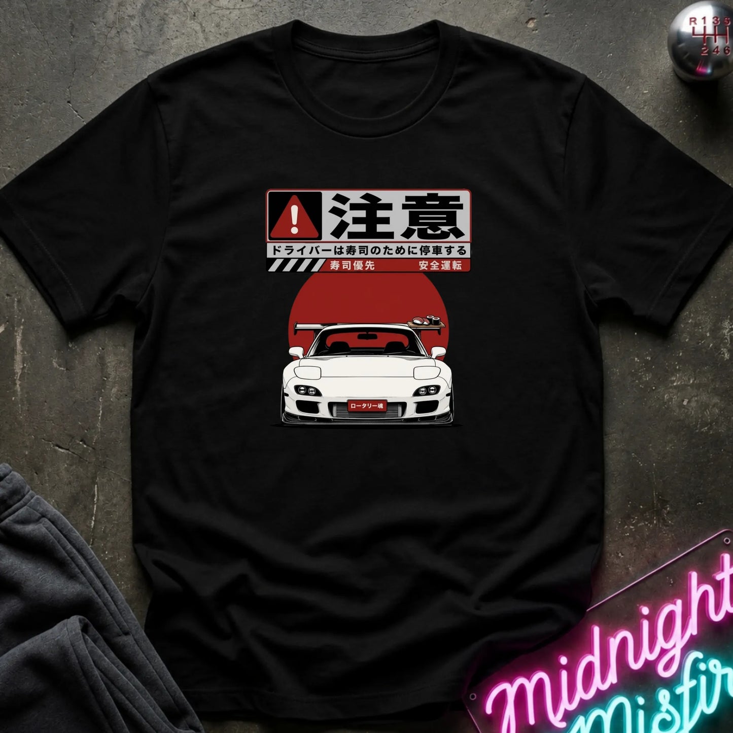 A black short-sleeve graphic T-shirt laid flat on a dark textured surface. The shirt features a Japanese warning sign design above a white sports car with a red sun graphic.